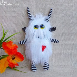 May include: A white furry monster toy with black and white striped arms and legs. The monster has large yellow eyes and a red heart on its chest.