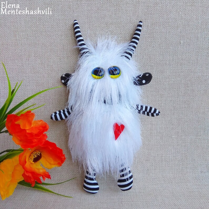 May include: A white furry monster toy with black and white striped arms and legs. The monster has large yellow eyes and a red heart on its chest.
