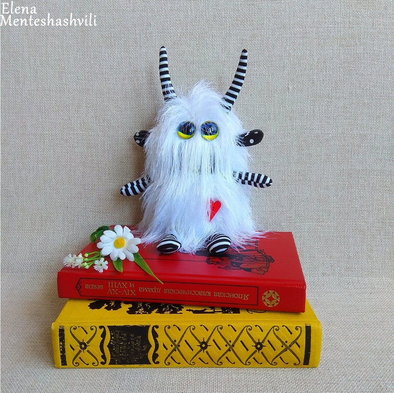 May include: A white furry monster toy with black and white striped arms and horns. The toy is sitting on top of two books, one red and one yellow. A white daisy is in front of the toy.