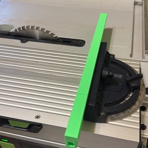 May include: A green and black table saw with a protractor attached to the side. The protractor is marked in degrees from 0 to 90. The table saw is on a white surface.