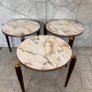 May include: Three small round side tables. Each table has a marble-look top with brown and beige veining, a gold-colored trim, and dark brown legs. The tables are arranged in a group.