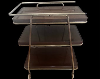 May include: A three-tiered serving cart with a chrome frame and brown glass shelves. The cart has wheels and is perfect for serving drinks and snacks.