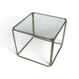 May include: A square side table with a clear glass top and a gold-colored metal frame. The table has a minimalist design and is suitable for various interior styles. The table measures approximately 18 inches in height and width.