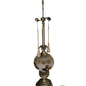 May include: A tall, gold-toned table lamp with a textured glass shade and a decorative finial. The lamp has a round base with a geometric design and a cord.