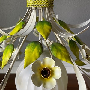 May include: White metal chandelier with green leaves and white flowers. The chandelier has a white globe shade.