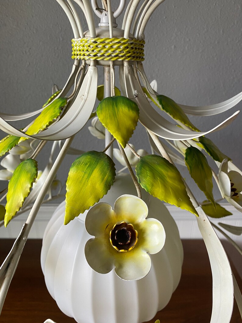 May include: White metal chandelier with green leaves and white flowers. The chandelier has a white globe shade.