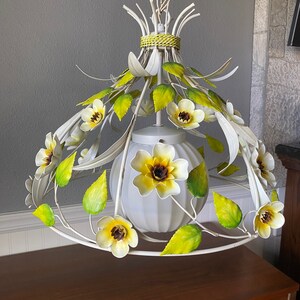 May include: White metal chandelier with yellow and white flowers and green leaves. The chandelier has a white globe shade.