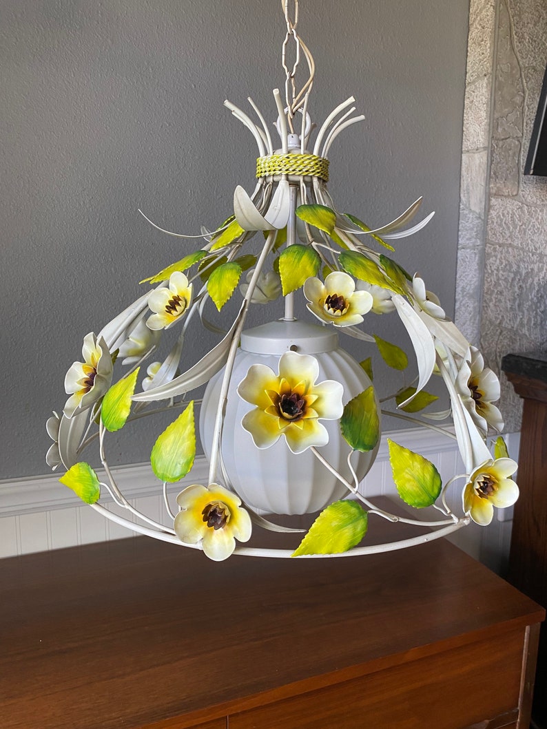 May include: White metal chandelier with yellow and white flowers and green leaves. The chandelier has a white globe shade.