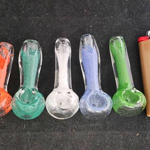 May include: Six glass pipes with different colored glass, including orange, green, white, blue, and green. The pipes are arranged in a row on a black surface. A brown lighter is to the right of the pipes.