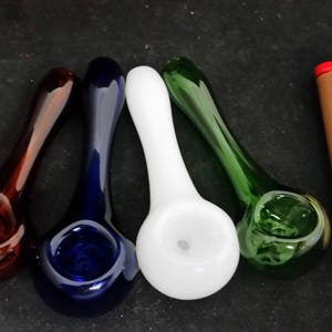May include: Four glass pipes in shades of red, blue, white, and green are arranged next to a brown lighter with a red top. The pipes have a spoon-like shape with a bowl and a long stem.