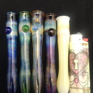 May include: Six glass smoking pipes with iridescent finishes. The pipes are arranged in a row from left to right. The pipes have different colored accents at the top. A lighter with a pink heart design is on the right side of the image.
