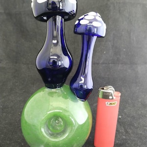 May include: A green glass water pipe with two blue glass mushroom-shaped bowls. The larger bowl has white polka dots. The smaller bowl has white polka dots. A red lighter is in the background.