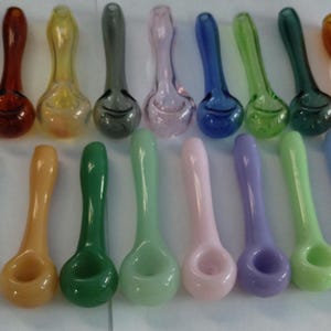 May include: A collection of 20 glass smoking pipes in various colors, including blue, green, white, and orange. The pipes are arranged in two rows, with a lighter in the background.