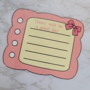 May include: A pink and cream-colored decorative notepad with the text "Today will be a good day." The notepad has a scalloped edge, a pink bow, and a series of horizontal lines for writing. There are four white circles on the left side.
