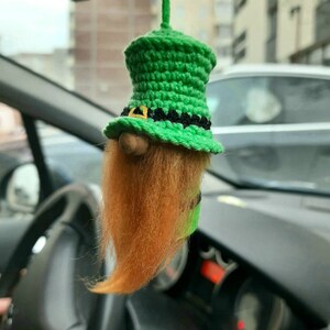 Könnte beinhalten: Ein grüner, gehäkelter Leprechaun-Hut mit einem schwarzen Band und einem langen, orangefarbenen Bart. Der Hut hängt an einem Rückspiegel in einem Auto.