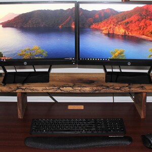 May include: A wooden monitor stand with a natural wood finish and black metal legs. Two black computer monitors are on top of the stand. A black keyboard and mouse are on a brown desk below the stand.