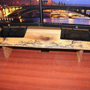 May include: A wooden monitor stand with a natural wood finish and black lightning bolt patterns. The stand has two triangular legs and is designed to hold two monitors.