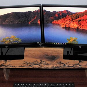 May include: Two black computer monitors with a scenic landscape wallpaper are mounted on a wooden stand with a natural wood grain finish.