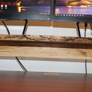 May include: A wooden monitor stand with a dark brown top and a lighter brown bottom shelf. The stand is designed to hold two monitors and has a space for cords to run through.