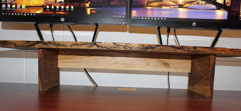 May include: A wooden monitor stand with a dark brown top and a lighter brown bottom shelf. The stand is designed to hold two monitors and has a space for cords to run through.