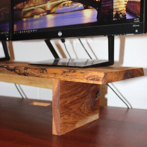 May include: A wooden monitor stand with a natural wood finish. The stand is made of a single piece of wood with a live edge. The stand is designed to hold two monitors and has a small shelf for a laptop or other items. The stand is sitting on a dark wood desk.