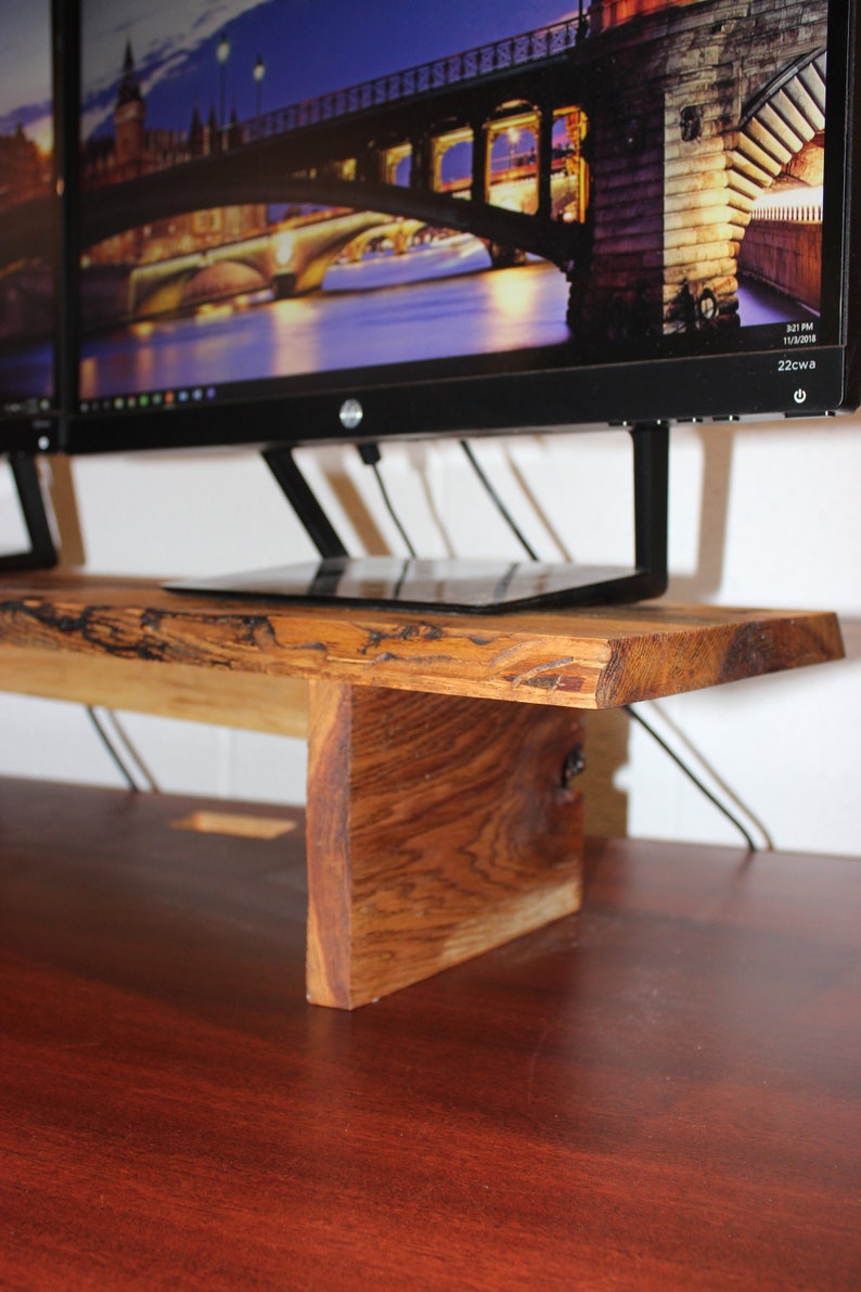 May include: A wooden monitor stand with a natural wood finish. The stand is made of a single piece of wood with a live edge. The stand is designed to hold two monitors and has a small shelf for a laptop or other items. The stand is sitting on a dark wood desk.