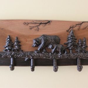 May include: A wooden wall-mounted key holder with a black and brown finish. The key holder features a cast iron design of a mother bear and cub walking through a forest with trees. There are five hooks for hanging keys.