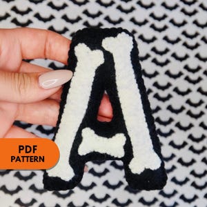 May include: A white felt letter A shaped like a bone, with a black felt background. The letter is being held by a hand. The text "PDF PATTERN" is visible in the lower left corner of the image.