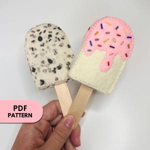 May include: Two felt popsicles on wooden sticks. One popsicle is white with black and brown sprinkles. The other popsicle is white with pink dripping icing and colorful sprinkles.