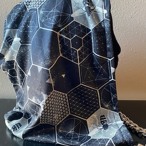 May include: A black and white patterned hood with a geometric design and bee illustrations. The hood is made of a soft, flowing fabric and features a silver chain detail. The design includes hexagons, lines, and starbursts.