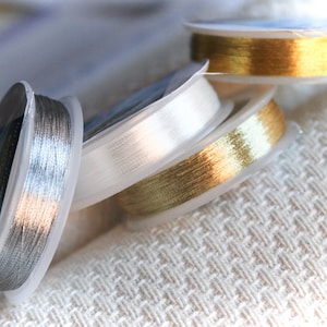 May include: Four spools of metallic thread in silver, white, gold, and a darker gold. The spools are stacked on top of each other on a white textured surface.
