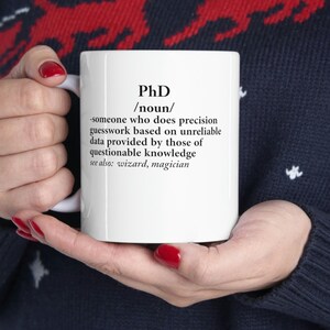 Könnte beinhalten: Eine weiße Kaffeetasse mit einer schwarz-weißen Definition eines Doktortitels. Die Definition lautet: "Jemand, der präzise Vermutungen anstellt, die auf unzuverlässigen Daten beruhen, die von Personen mit fragwürdigem Wissen geliefert werden. Siehe auch: Zauberer, Magier."