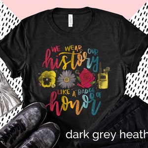 May include: Dark grey heather t-shirt with the text "We wear our history like a badge of honor" in rainbow colors. The text is surrounded by flowers and a walkie-talkie.
