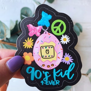 May include: A black and white sticker with a pink and yellow Tamagotchi, a peace sign, butterflies, and flowers. The text reads "90's kid 4ever".
