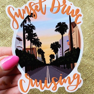 May include: A white sticker with orange text that reads "Sunset Drive Cruising" with a sunset scene of palm trees and a road in the background.