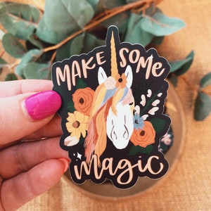 May include: A black sticker with a white unicorn with a rainbow mane and tail. The unicorn is surrounded by orange, yellow, and blue flowers. The text "Make some magic" is written in white cursive lettering.