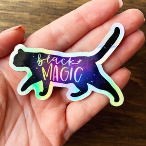 May include: A holographic sticker of a black cat with a galaxy background and the text "black magic" in white.