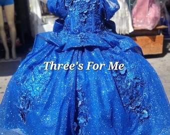 May include: A blue sparkly dress with a tulle skirt and floral embellishments. The dress has a sweetheart neckline and a fitted bodice. The dress is on a mannequin and the text "Three's For Me" is visible on the skirt.