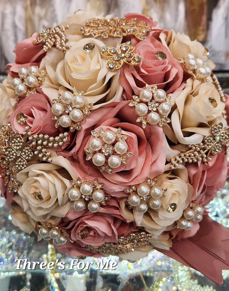 May include: A bridal bouquet made of blush pink and ivory roses with gold and pearl embellishments. The bouquet is adorned with gold brooches and pearl accents. "Three's For Me" is written on the bottom of the image.