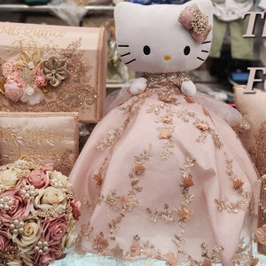 May include: A pink and gold themed quincea&ntilde;era decoration set with a Hello Kitty doll wearing a pink dress, a pillow with the text "Mis Quince A&ntilde;os", and several boxes and books with the same text. The text "Three's For Me" is written above the pillow.