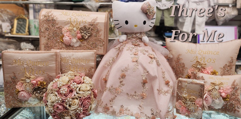 May include: A pink and gold themed quincea&ntilde;era decoration set with a Hello Kitty doll wearing a pink dress, a pillow with the text "Mis Quince A&ntilde;os", and several boxes and books with the same text. The text "Three's For Me" is written above the pillow.