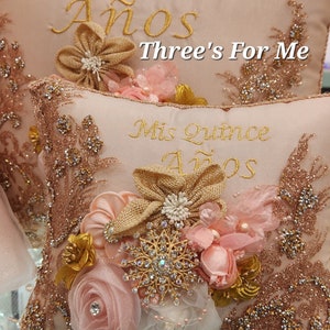 May include: Two pink and gold decorative pillows with the text "Mis Quince A&ntilde;os" and "Three's For Me" embroidered on them. The pillows are decorated with pink and gold fabric flowers and rhinestones.