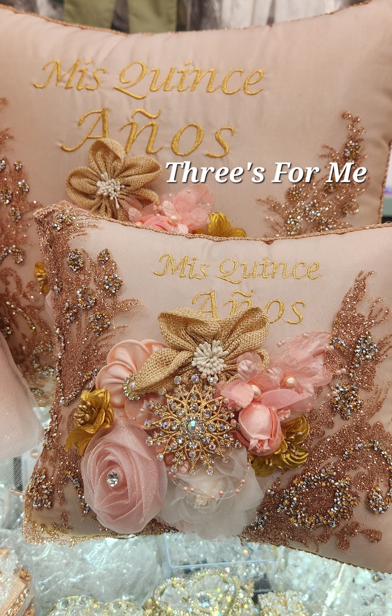 May include: Two pink and gold decorative pillows with the text "Mis Quince A&ntilde;os" and "Three's For Me" embroidered on them. The pillows are decorated with pink and gold fabric flowers and rhinestones.