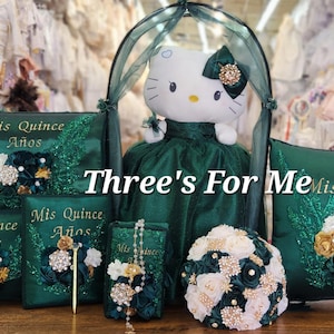 May include: A green and gold quinceañera pillow set with a Hello Kitty doll wearing a green dress. The set includes three pillows, a bouquet, and a small book. The pillows are embroidered with the text "Mis Quince Años".