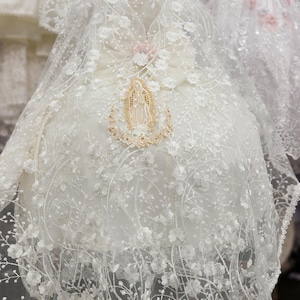 May include: White lace christening gown with floral embroidery and a decorative religious emblem. The gown features a high neckline and a flowing skirt, with the text "Estilo #4 Back" at the top.