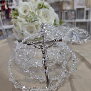 May include: A religious rosary featuring clear, faceted crystal beads interspersed with smooth, frosted white oval beads. The rosary includes a silver-toned metal crucifix with a detailed figure of Jesus and the letters INRI inscribed at the top.