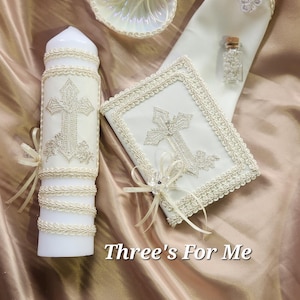 May include: A set of three white baptism items with a cross design. The set includes a candle, a small book, and a small bottle. The items are decorated with white lace and pearls. The text "Three's For Me" is visible in the image.