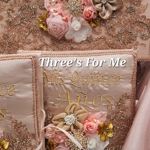 May include: Three pink satin boxes with gold embroidery that reads "Mis Quince A&ntilde;os" and "Three's For Me". Each box is decorated with a pink and gold flower arrangement.