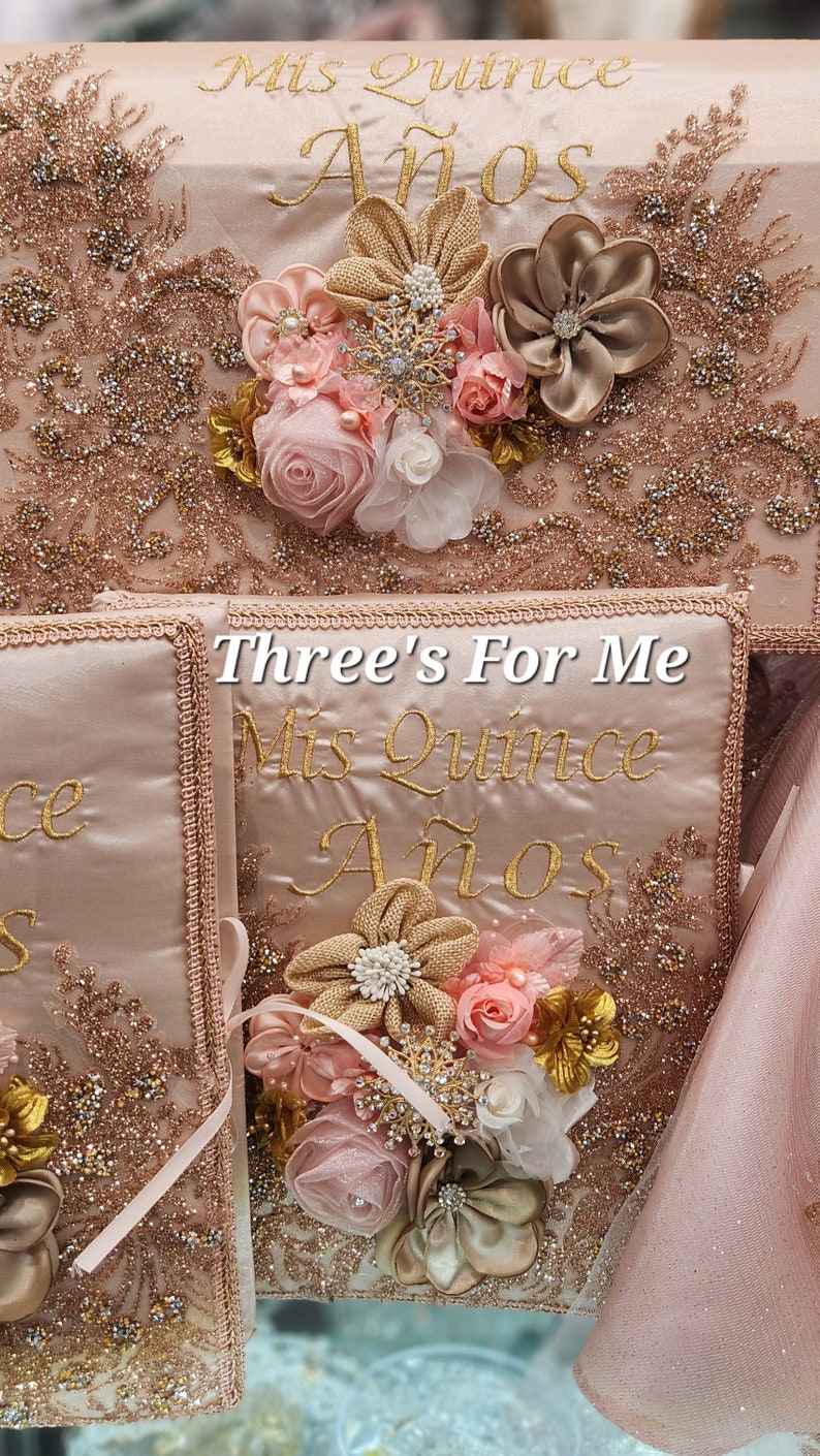 May include: Three pink satin boxes with gold embroidery that reads "Mis Quince A&ntilde;os" and "Three's For Me". Each box is decorated with a pink and gold flower arrangement.