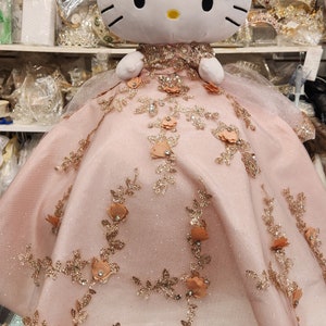 May include: A plush Hello Kitty doll wearing a pink dress with gold glitter and floral embellishments.